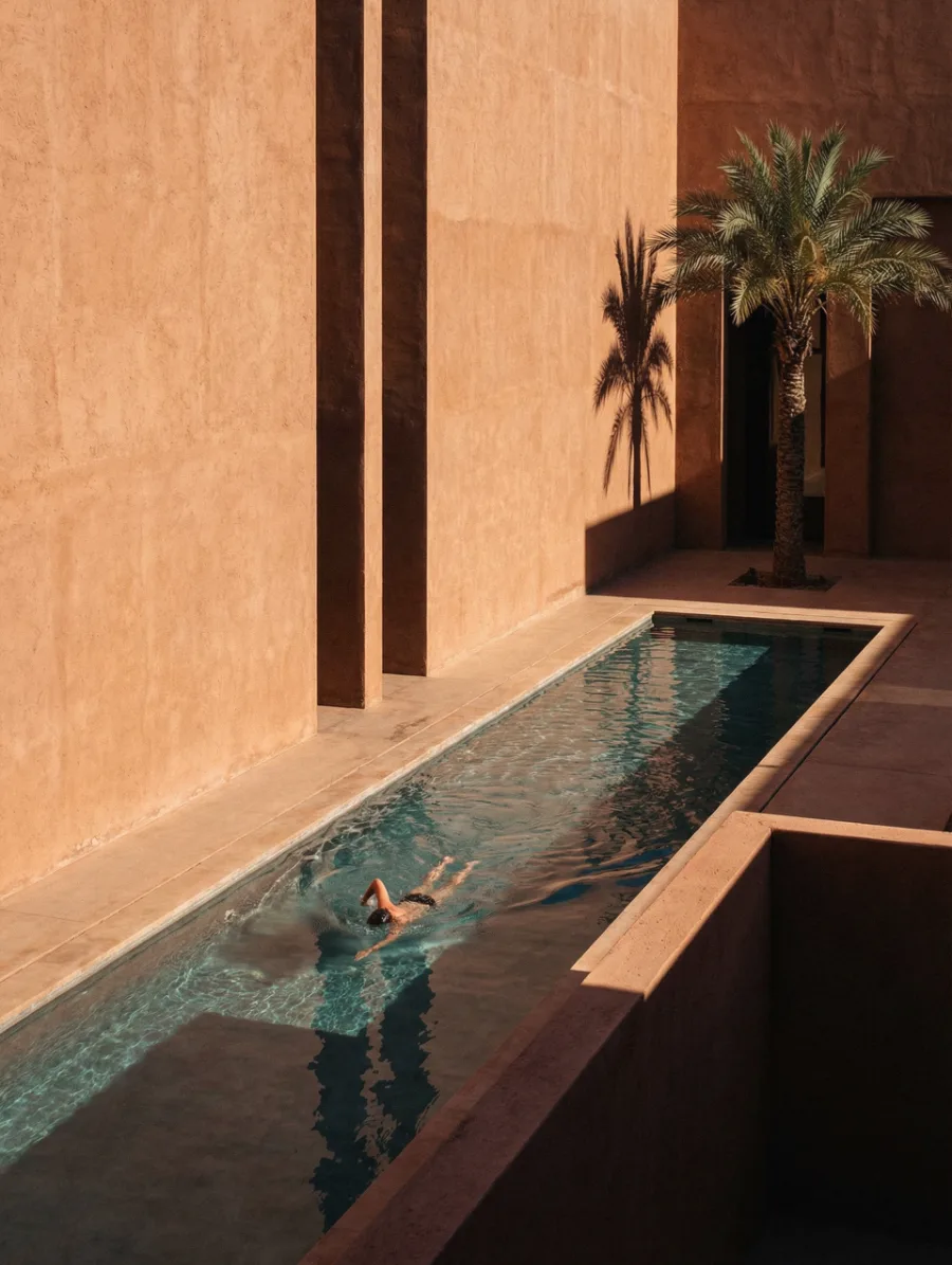 A swimmer crossing a long narrow pool between sunlit ochre concrete walls.